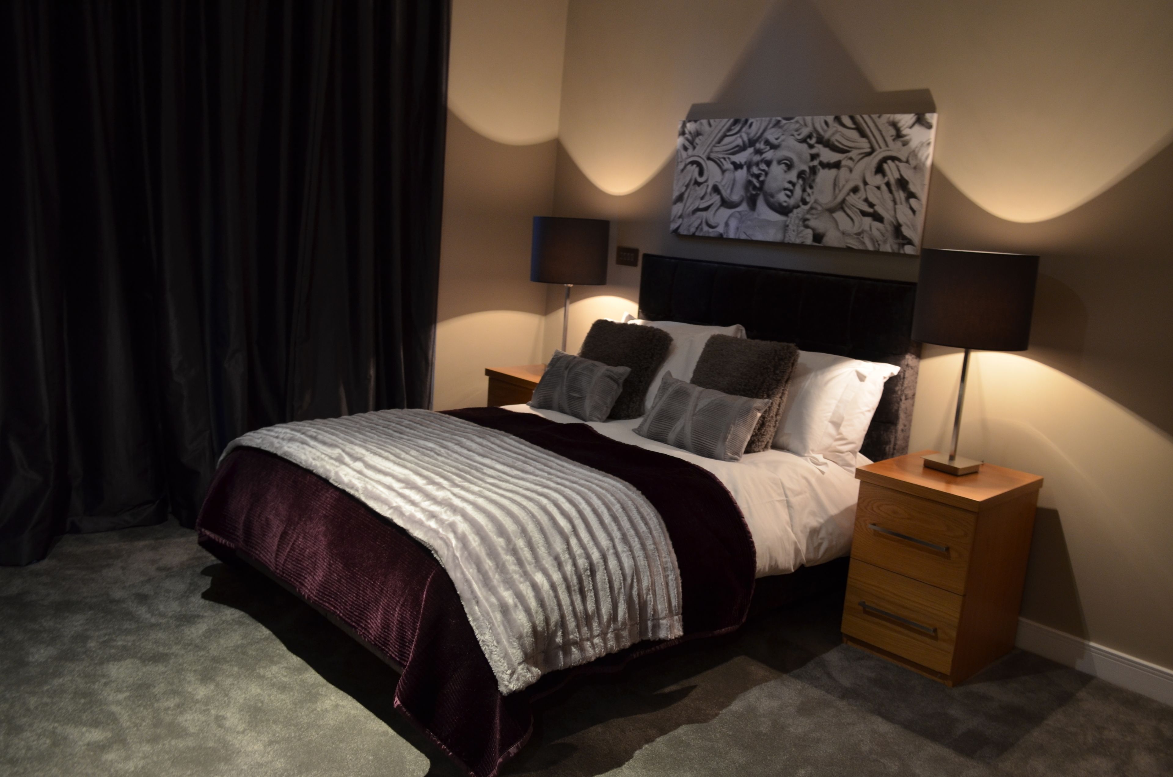 A bedroom with a bed, two bedside tables, a nightstand, a large abstract artwork above the bed, and a dark curtain.