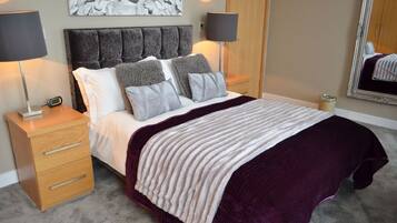 A bedroom with a bed, two bedside tables, a nightstand, a lamp, a drawer unit, and a mirror.