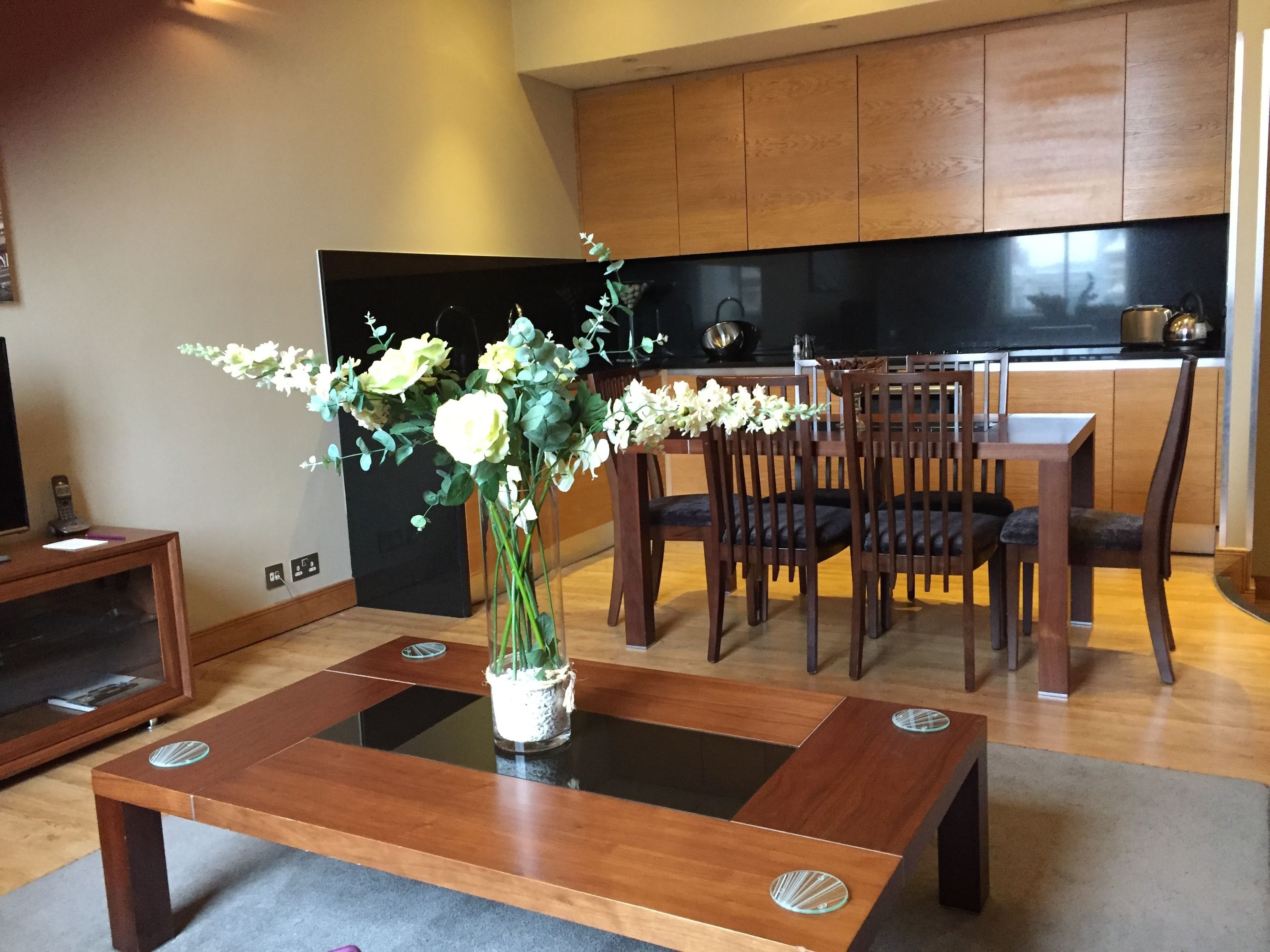 Una sala de estar moderna con una mesa de centro de madera, un florero con flores y un comedor con sillas y una mesa de madera.