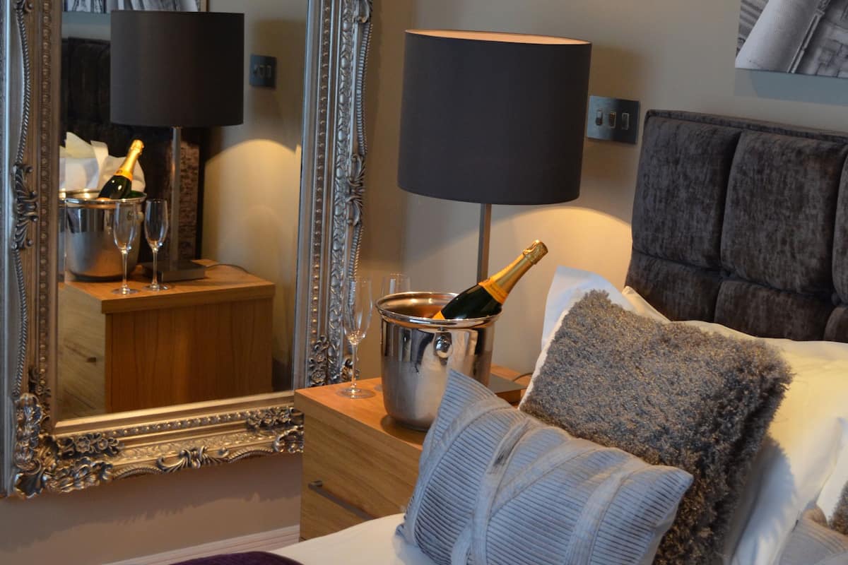 A hotel room with a large ornate mirror, a bed with multiple pillows, a bedside table with a lamp, and a bucket with champagne.