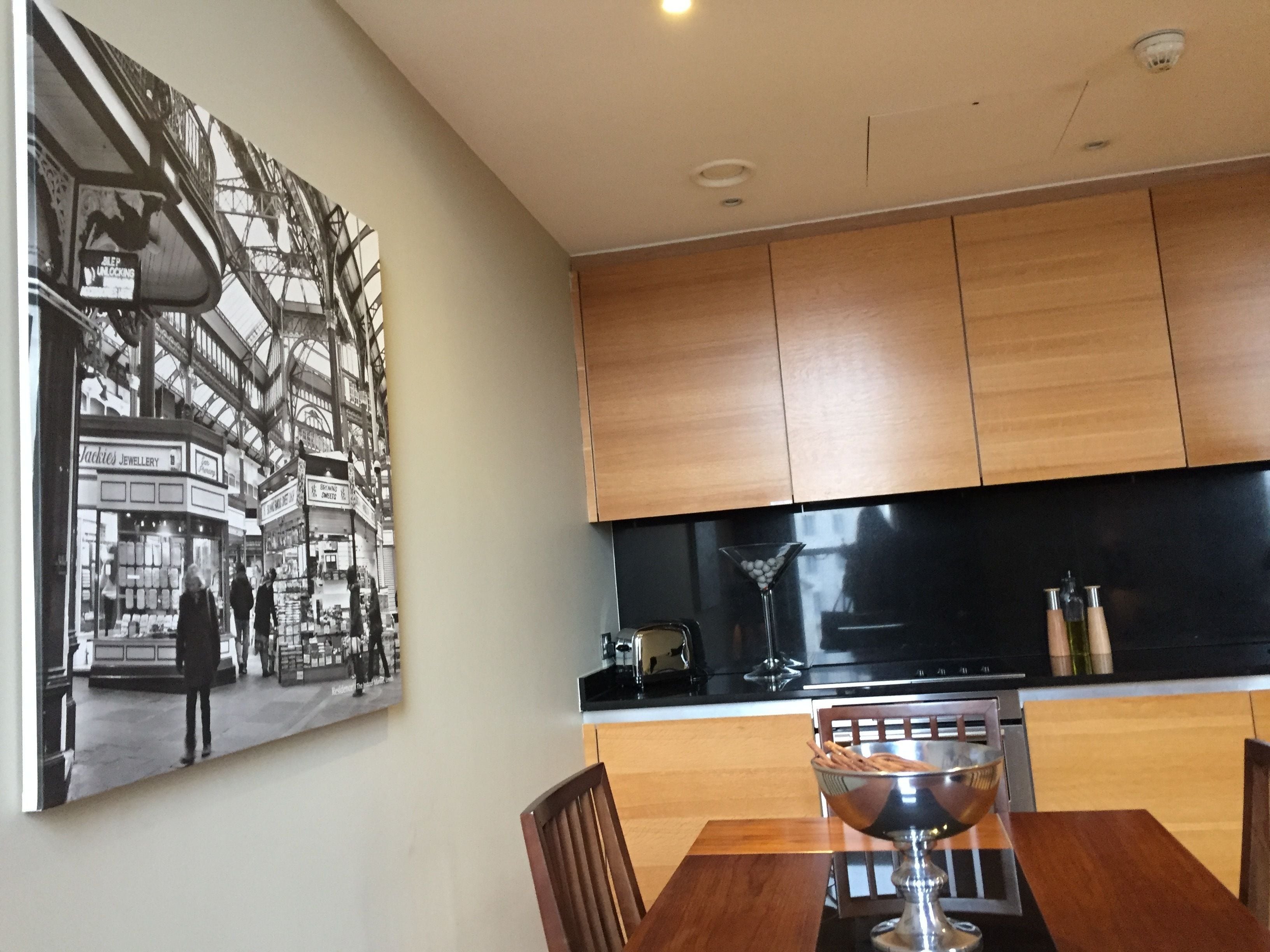 A modern kitchen with wooden cabinets, a black countertop, and a dining table with chairs. A large black and white photograph is displayed on the wall.