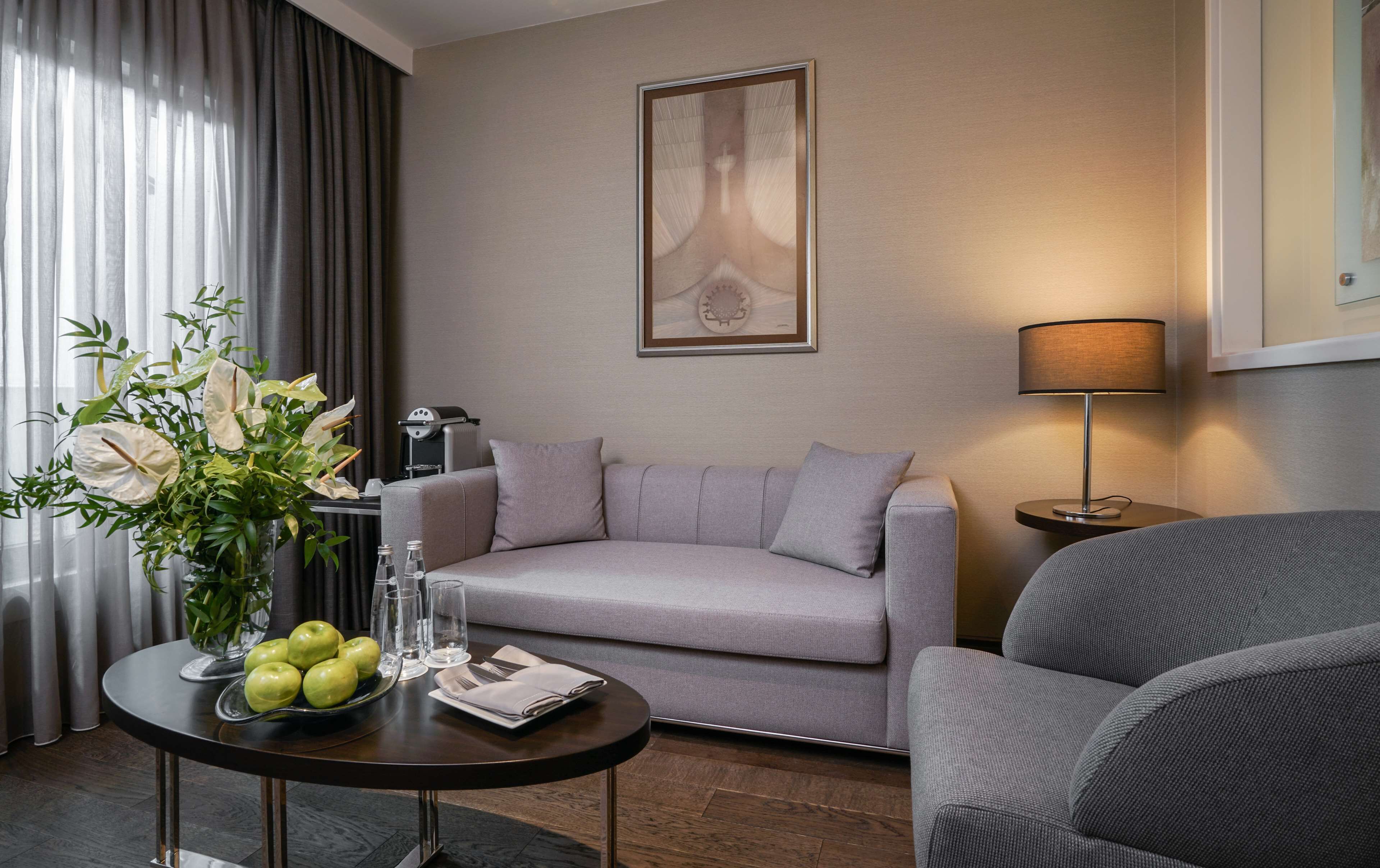 Una habitación de hotel moderna con un sofá gris, una mesa redonda con fruteros y vasos, una lámpara y una obra de arte enmarcada en la pared.