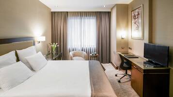 A hotel room with a large bed, a desk with a computer, a chair, a TV, and a window with curtains.