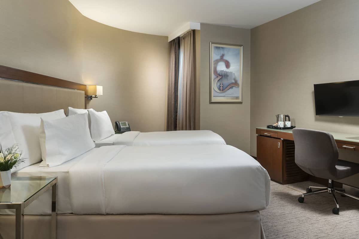 A hotel room with two beds, a desk, a chair, a flat-screen TV, a telephone, and a painting on the wall.