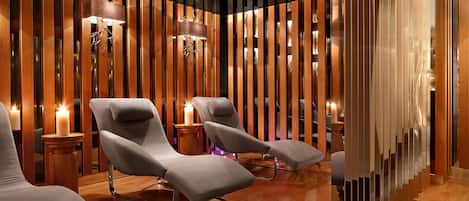 A modern spa area with wooden paneling, two grey lounge chairs, and a small wooden table with a candle.