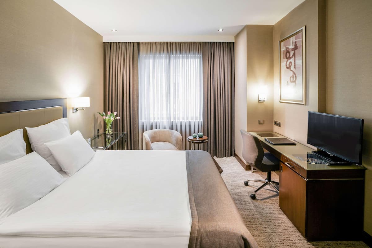 Une chambre d’hôtel comprenant un lit, un bureau avec un ordinateur, une chaise, une télévision, une lampe et une fenêtre avec des rideaux.