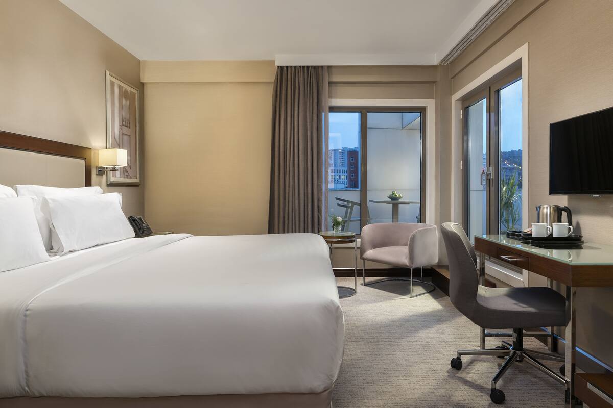 A hotel room with a large bed, a desk with a chair, a TV, and a window with a city view.