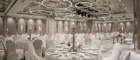 Un amplio salón de banquetes con mesas redondas, sillas blancas y candelabros elegantes.