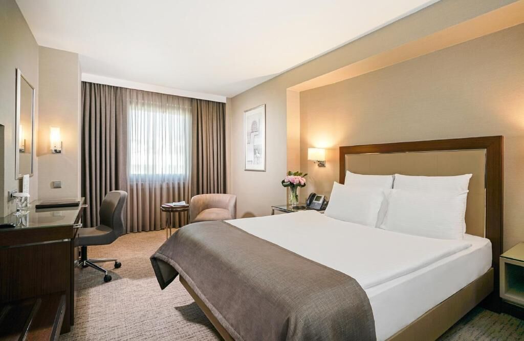 A hotel room with a large bed, a desk with a chair, a small table with a lamp, and a large window with curtains.