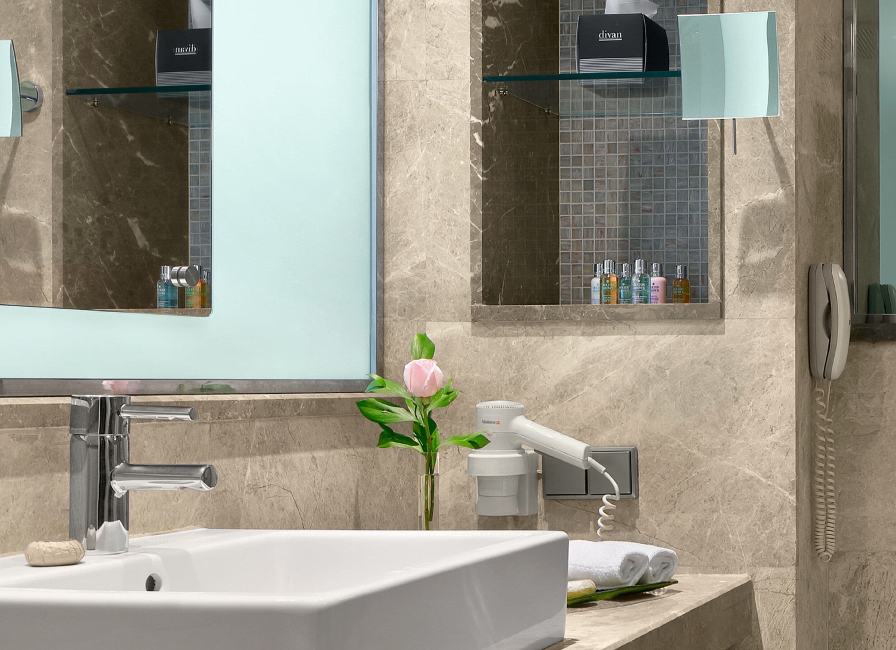 Un baño moderno con un espejo grande, un lavamanos blanco, un secador de pelo y una ventana pequeña con artículos de aseo.