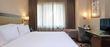 Habitación de hotel con una cama grande, un escritorio con silla, televisor y ventana con cortinas.