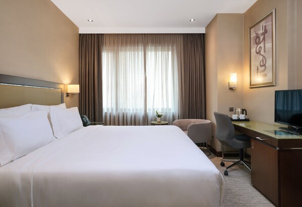 A hotel room with a large bed, a desk with a chair, a television, and a window with curtains.