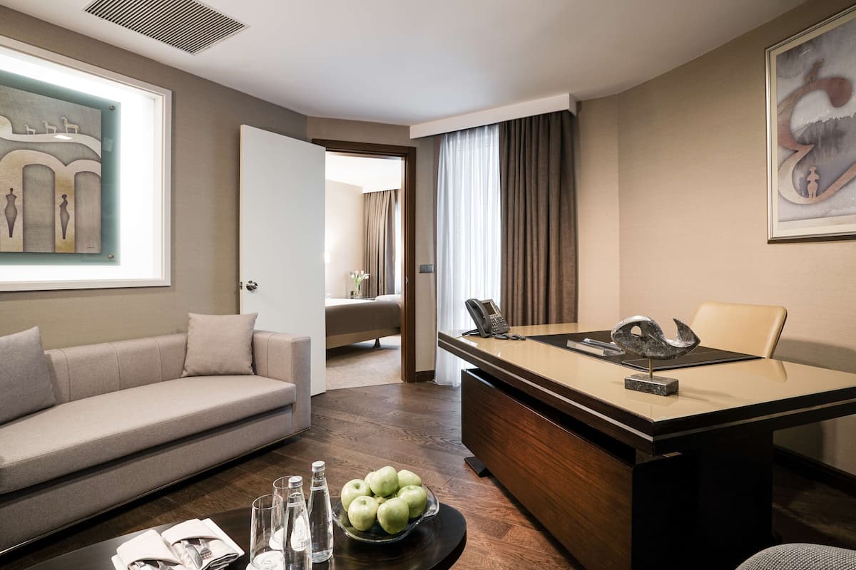 A modern hotel room with a sofa, a dining table with a vase of apples, a phone, and a painting on the wall.