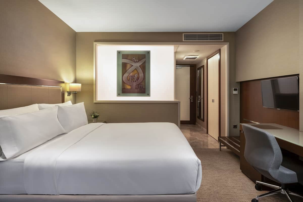 A hotel room with a large bed, a desk with a chair, a television, and a wall-mounted artwork.