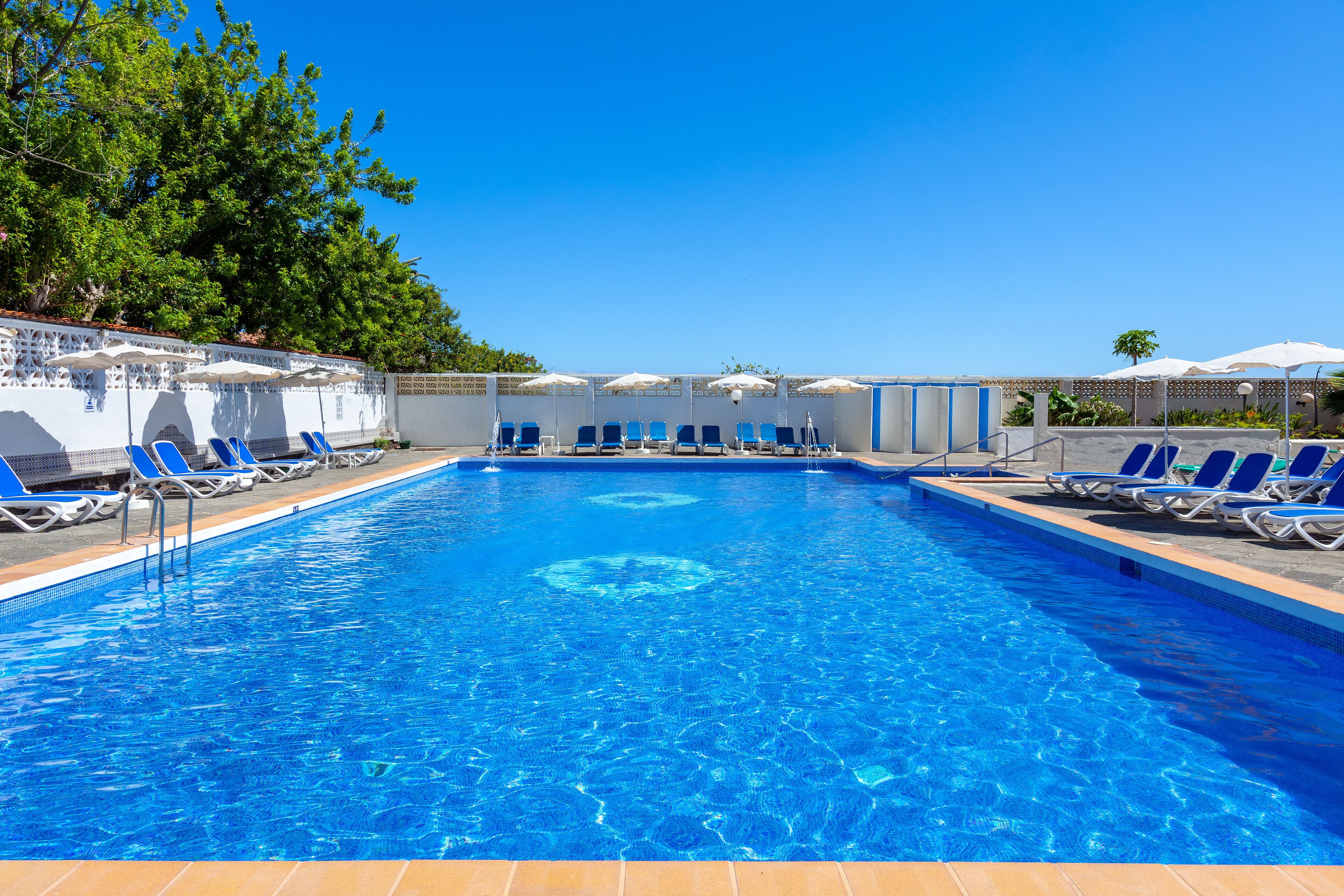Una piscina rectangular con agua azul cristalina, rodeada de sillas reclinables azules y sombrillas blancas.