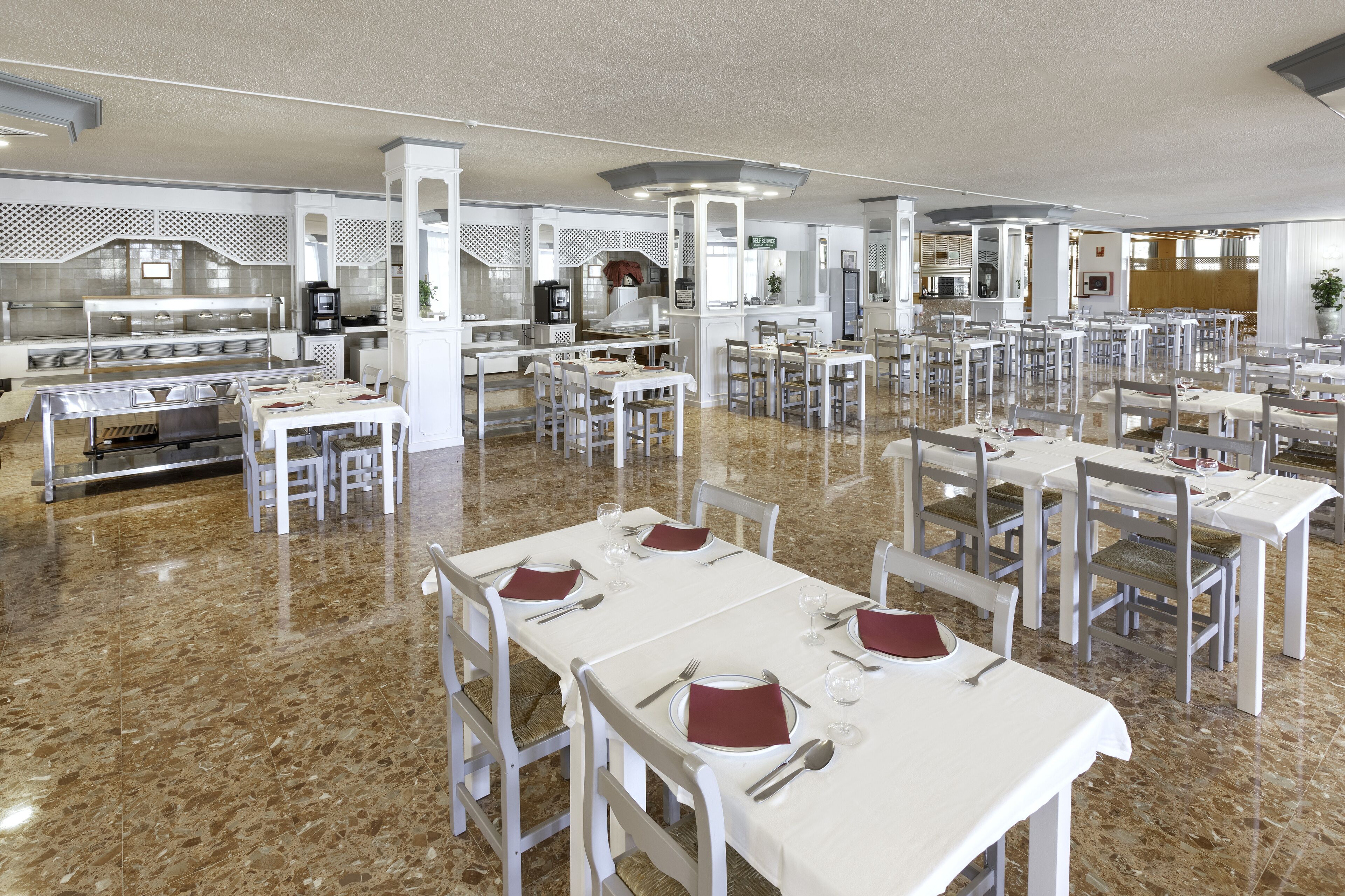 A spacious dining area with multiple tables and chairs, a buffet line, and a kitchen in the background.