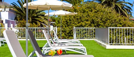 A row of white lounge chairs with umbrellas, arranged on a well-maintained lawn with a metal fence and palm trees in the background.