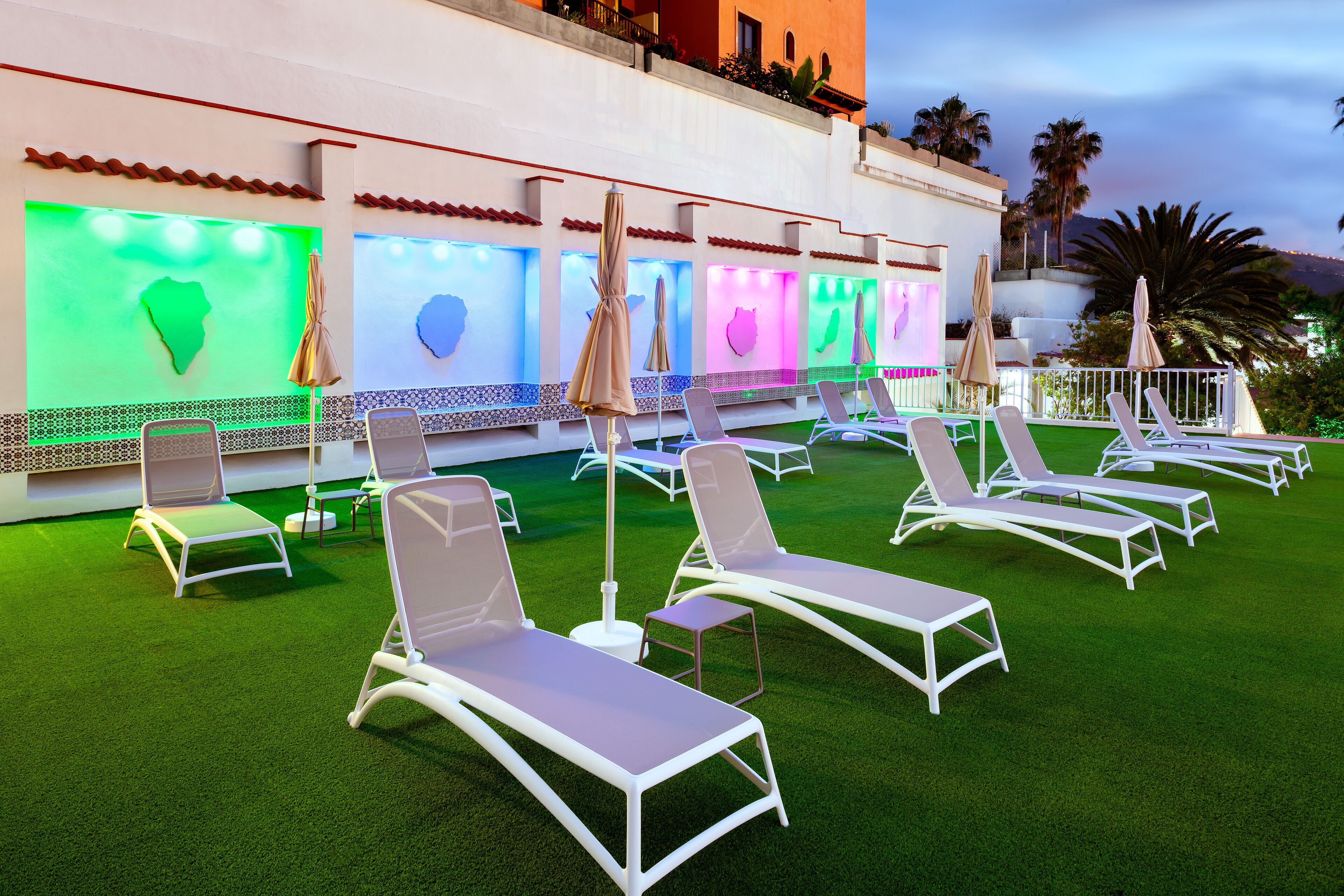 Un espace aménagé au bord de la piscine, avec des chaises longues blanches et des parasols, une pelouse artificielle verte et des éléments d’eau illuminés.