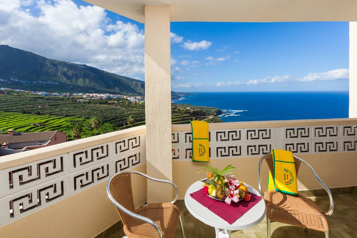 Un balcon avec vue sur l’océan et un paysage montagneux. On y trouve deux fauteuils en osier et une petite table sur laquelle sont posés un panier de fruits et une serviette jaune.