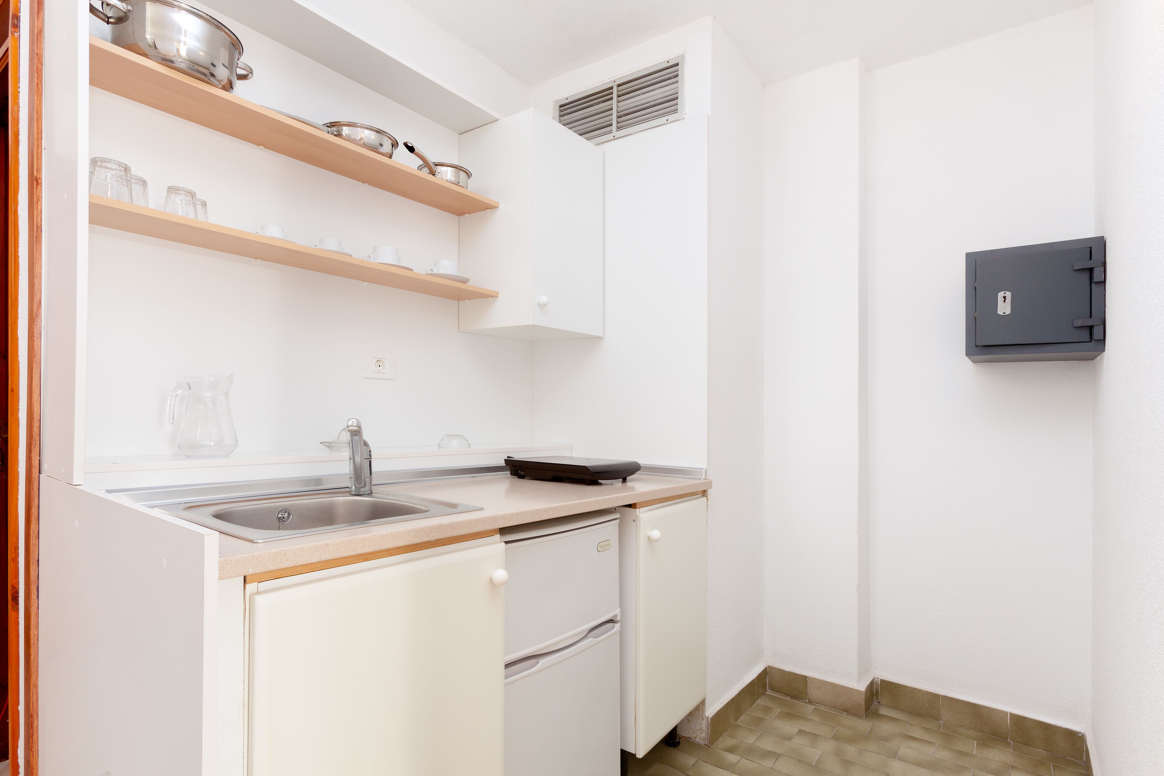 A compact kitchen with a sink, countertop, and built-in oven. There is a small refrigerator and a wall-mounted safe.