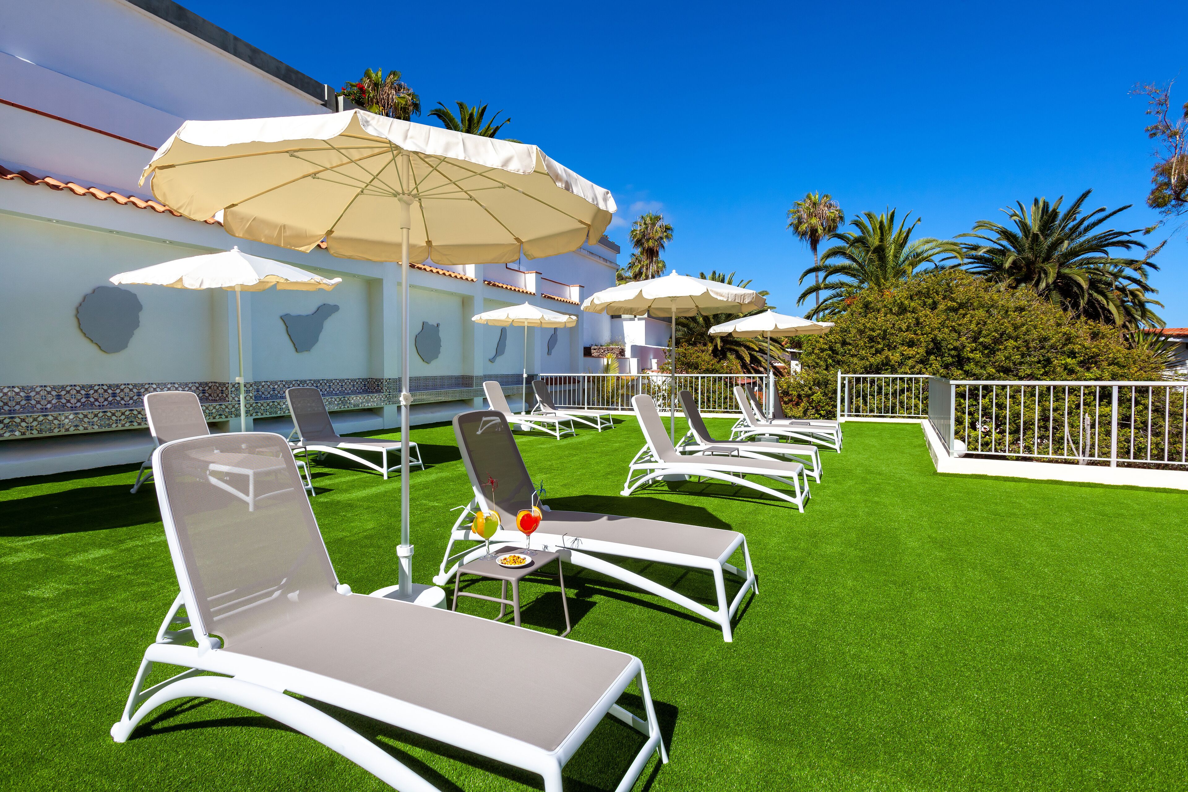 A rooftop lounge area with white sun loungers and beige umbrellas, surrounded by a white fence and palm trees.