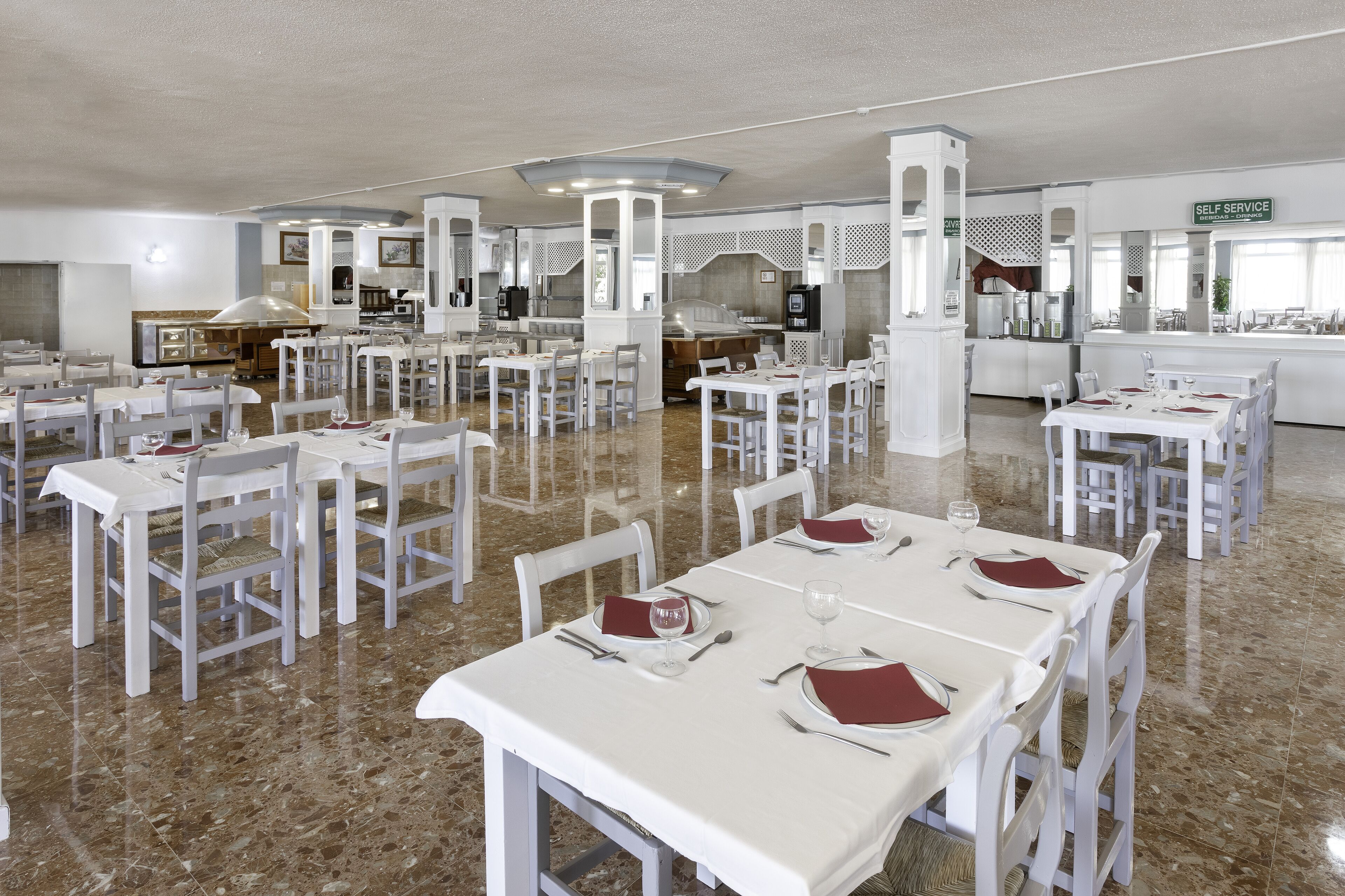 Un amplio comedor con varias mesas y sillas, una estación de autoservicio y una cocina al fondo.
