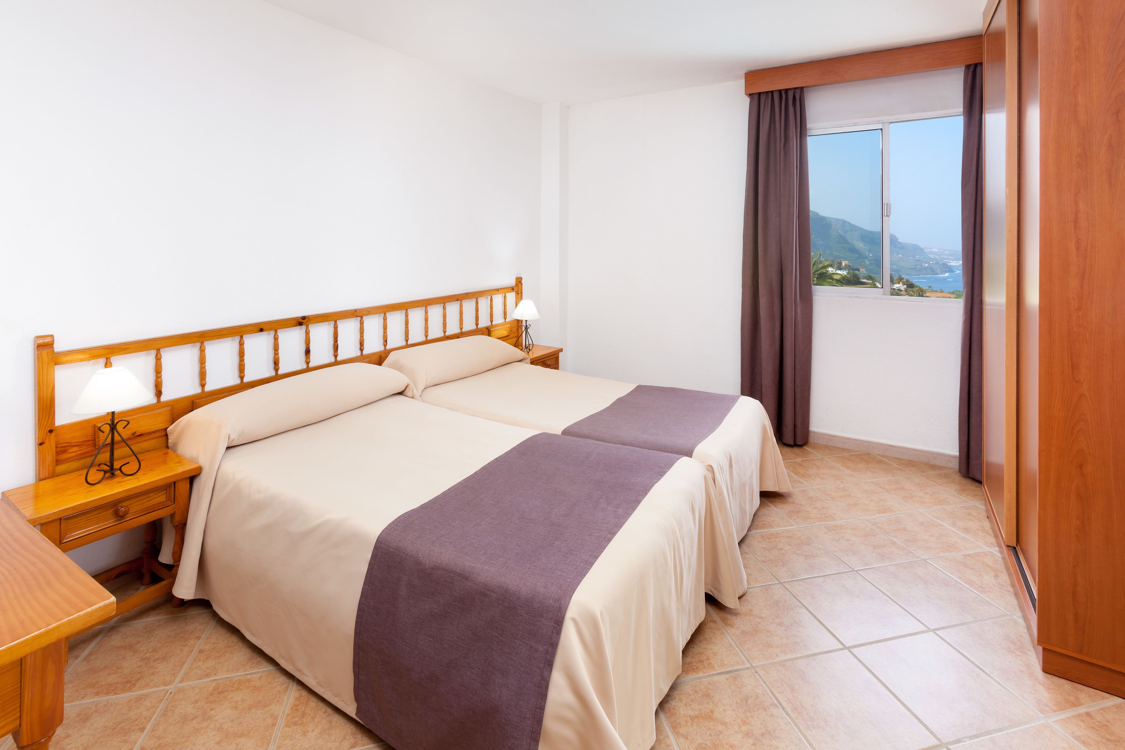 Cama doble con cabecera de madera, dos mesitas de noche con lámparas, vista al exterior por una ventana y un balcón con cortinas.
