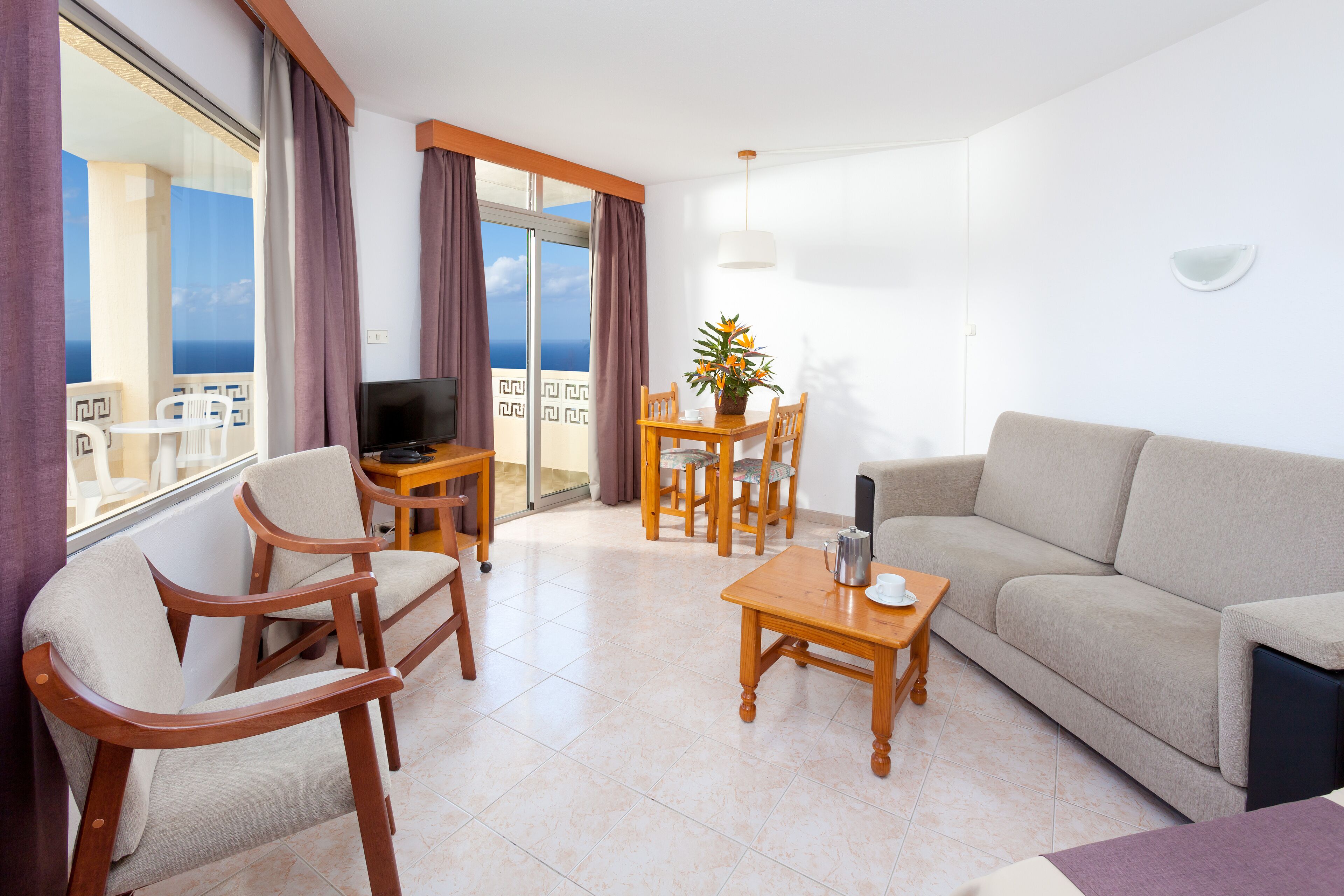 A modern living room with a sofa, chairs, a coffee table, and a TV. There is a balcony with a view of the sea.