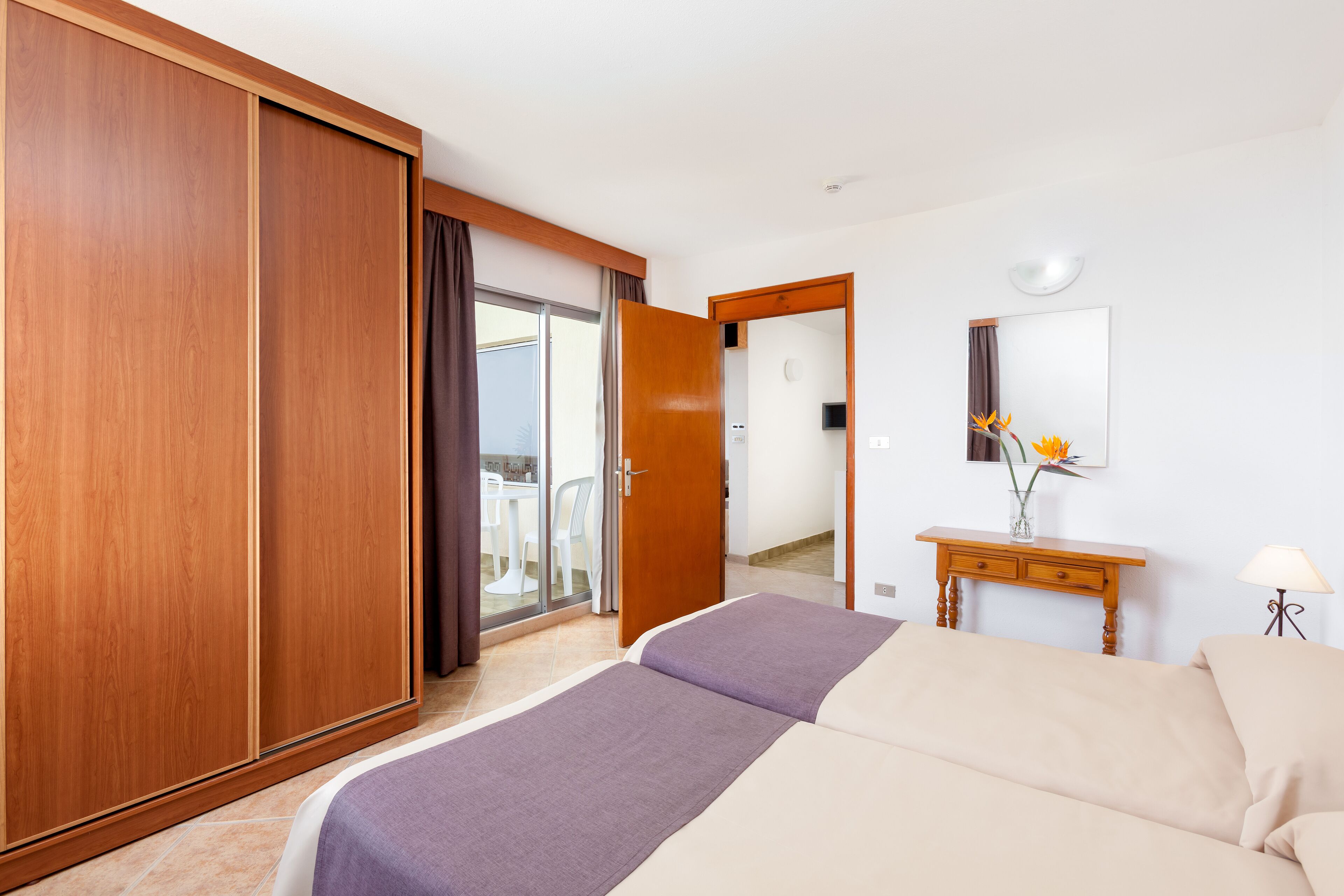 Een hotelkamer met een grote kledingkast, een bed met een paars dekbedovertrek, een houten nachtkastje met een lamp en uitzicht op het balkon door de deur.