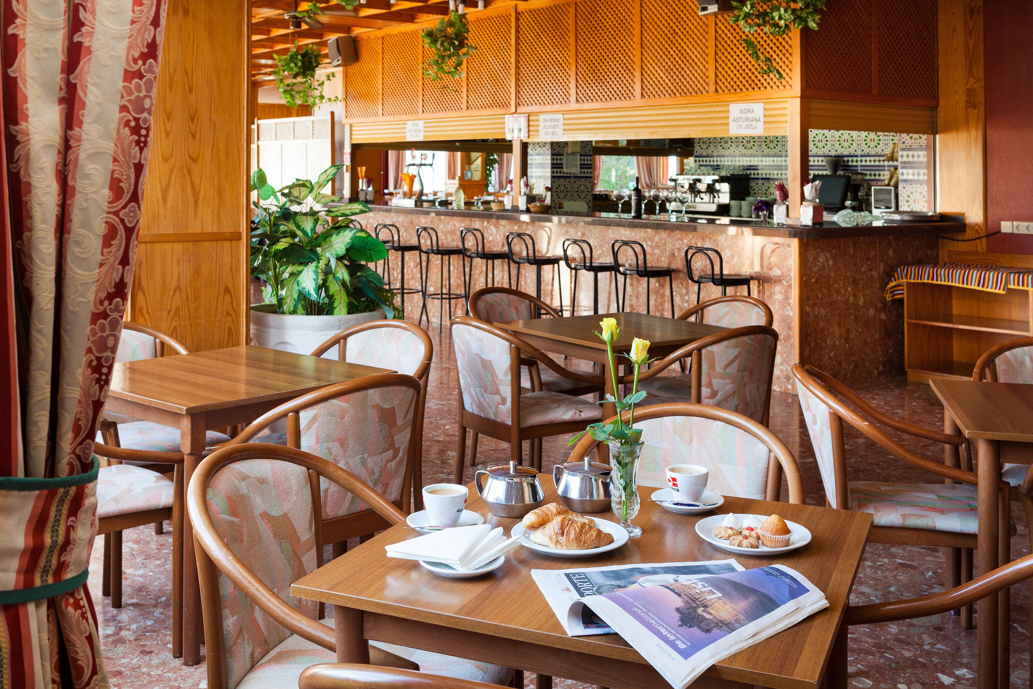 A café with wooden tables and chairs, a bar area, and potted plants.