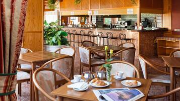 A café with wooden tables and chairs, a bar area, and potted plants.
