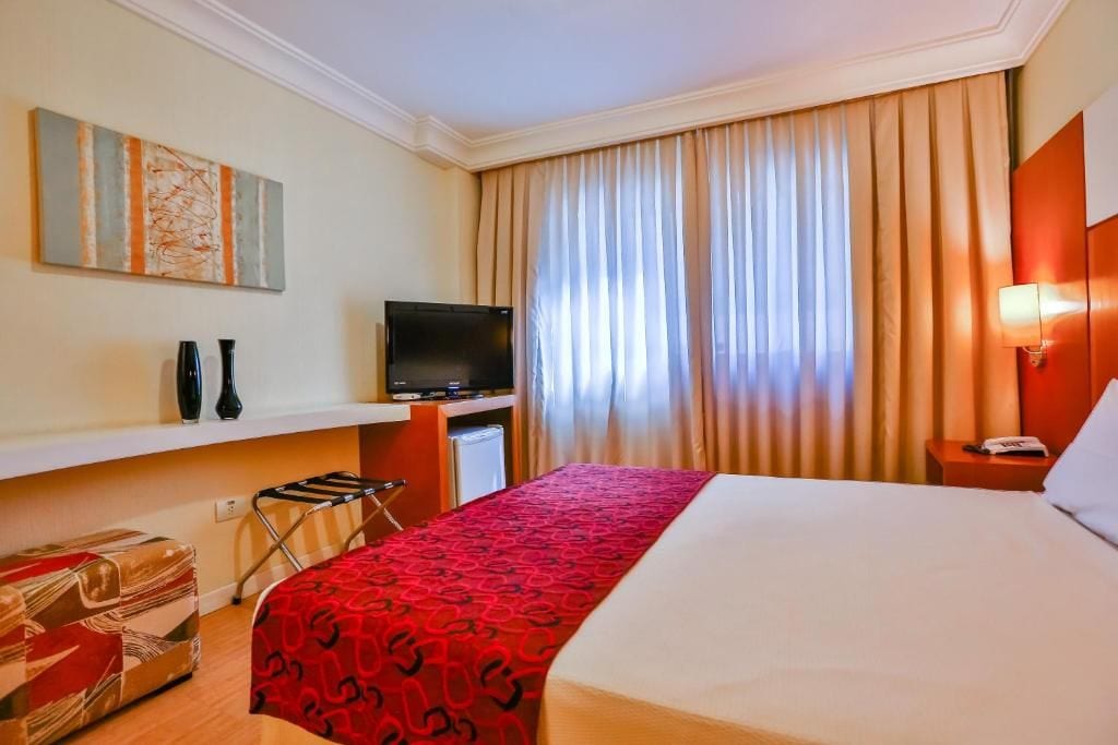 A hotel room with a bed, a TV, a desk, a chair, and a window with curtains.
