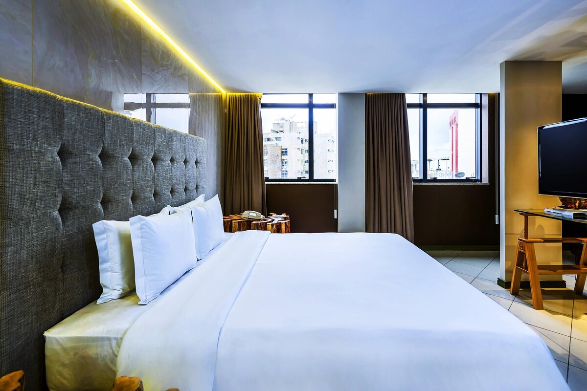 A hotel room with a large bed, a television, a desk, and a view of the city through the windows.