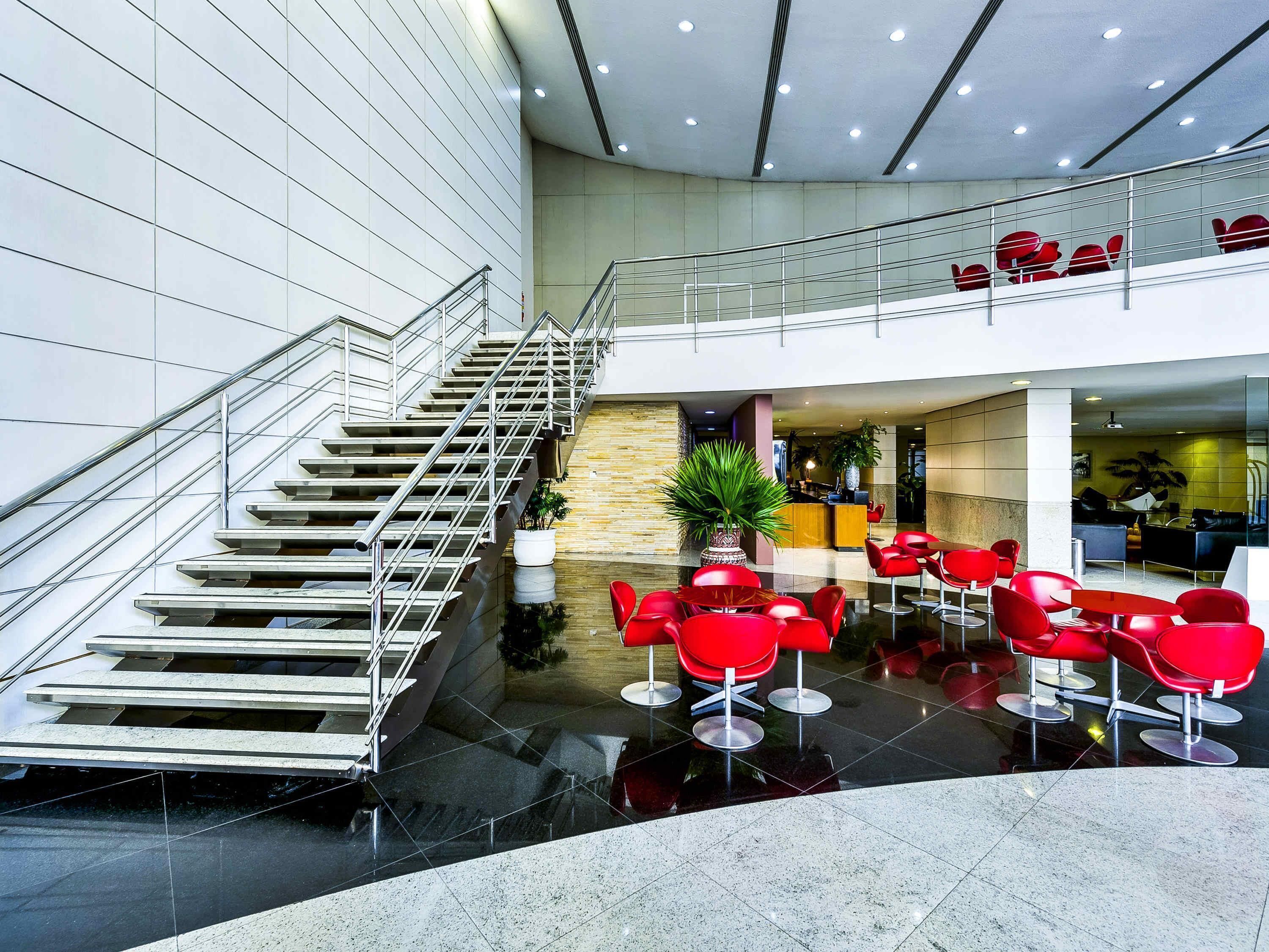 A modern hotel lobby with a central staircase, red seating area, and a reception desk.