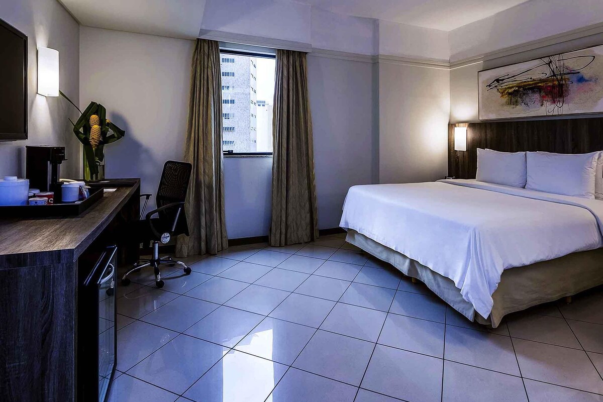 A hotel room with a bed, a desk with a coffee machine, a chair, a window with curtains, and a painting on the wall.