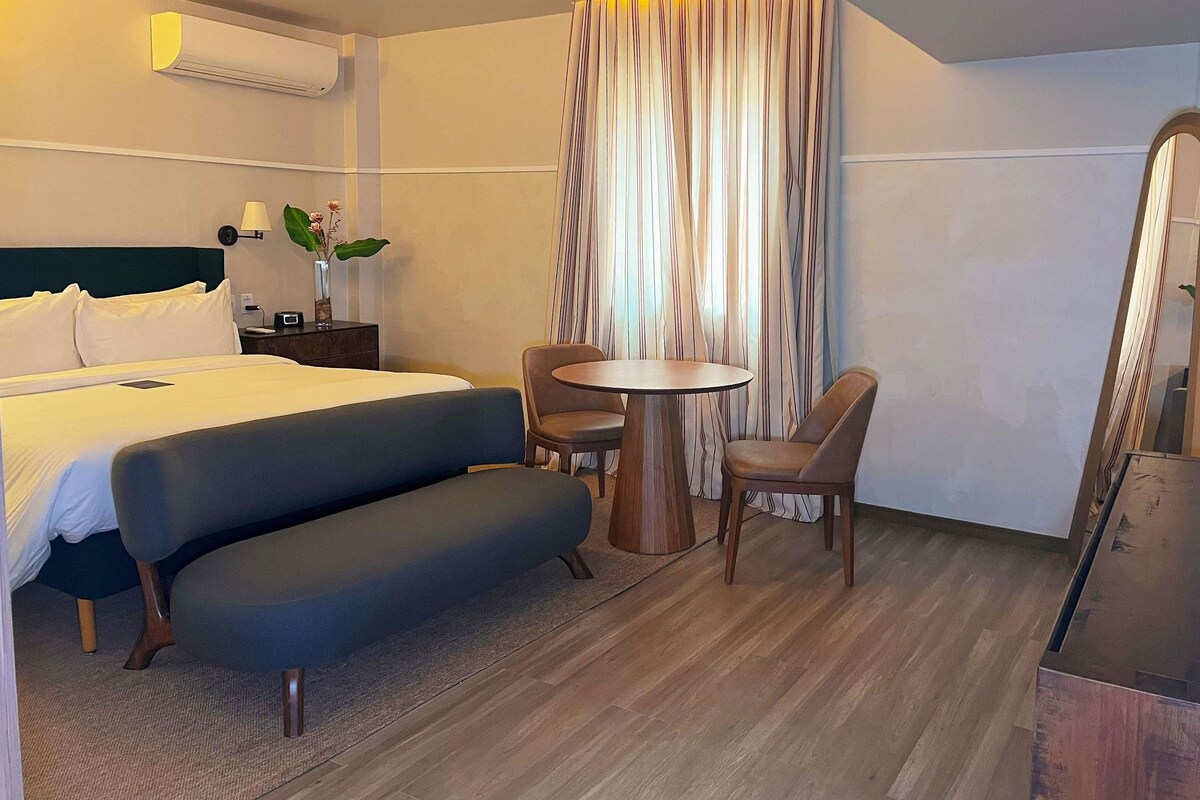 A hotel room with a bed, a sofa, a small table with chairs, a mirror, and a window with curtains.