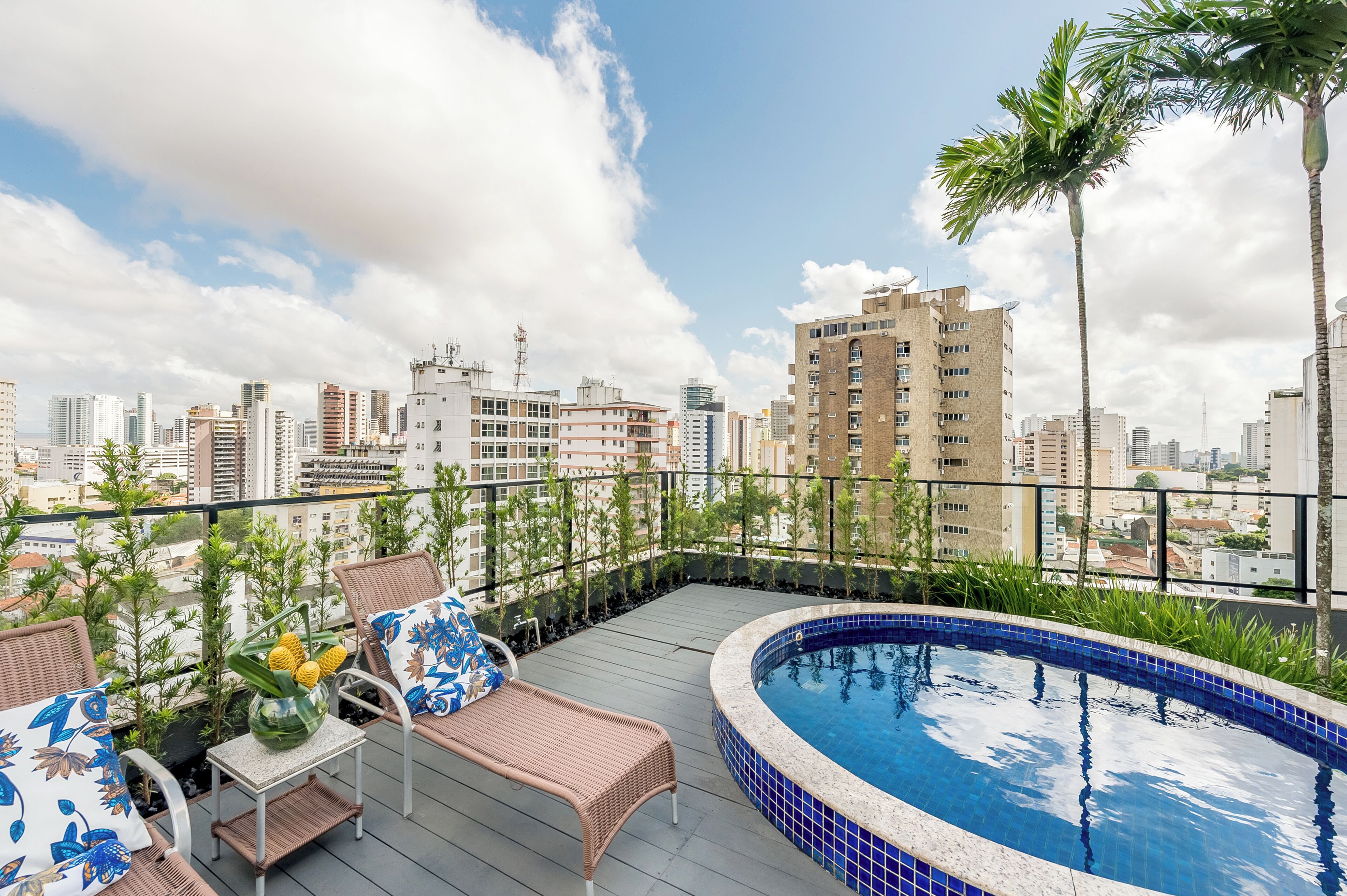 Una piscina en la azotea con sillones reclinables y una mesa, rodeada de plantas en macetas y con vistas a un paisaje urbano.