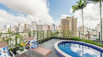Una piscina en la azotea con sillones reclinables y una mesa, rodeada de plantas en macetas y con vistas a un paisaje urbano.