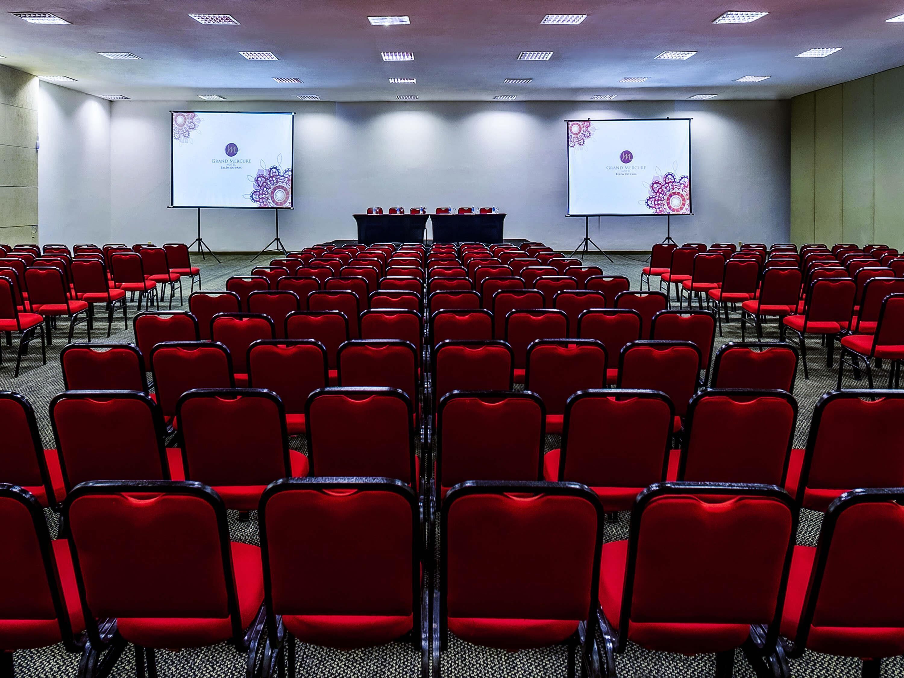 A conference room with rows of red chairs, two projection screens, and a podium at the front.