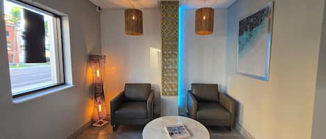 Un espace d’attente moderne comprenant deux fauteuils en cuir, une table ronde en marbre avec un magazine, un grand lampadaire et une grande fenêtre donnant sur la rue.