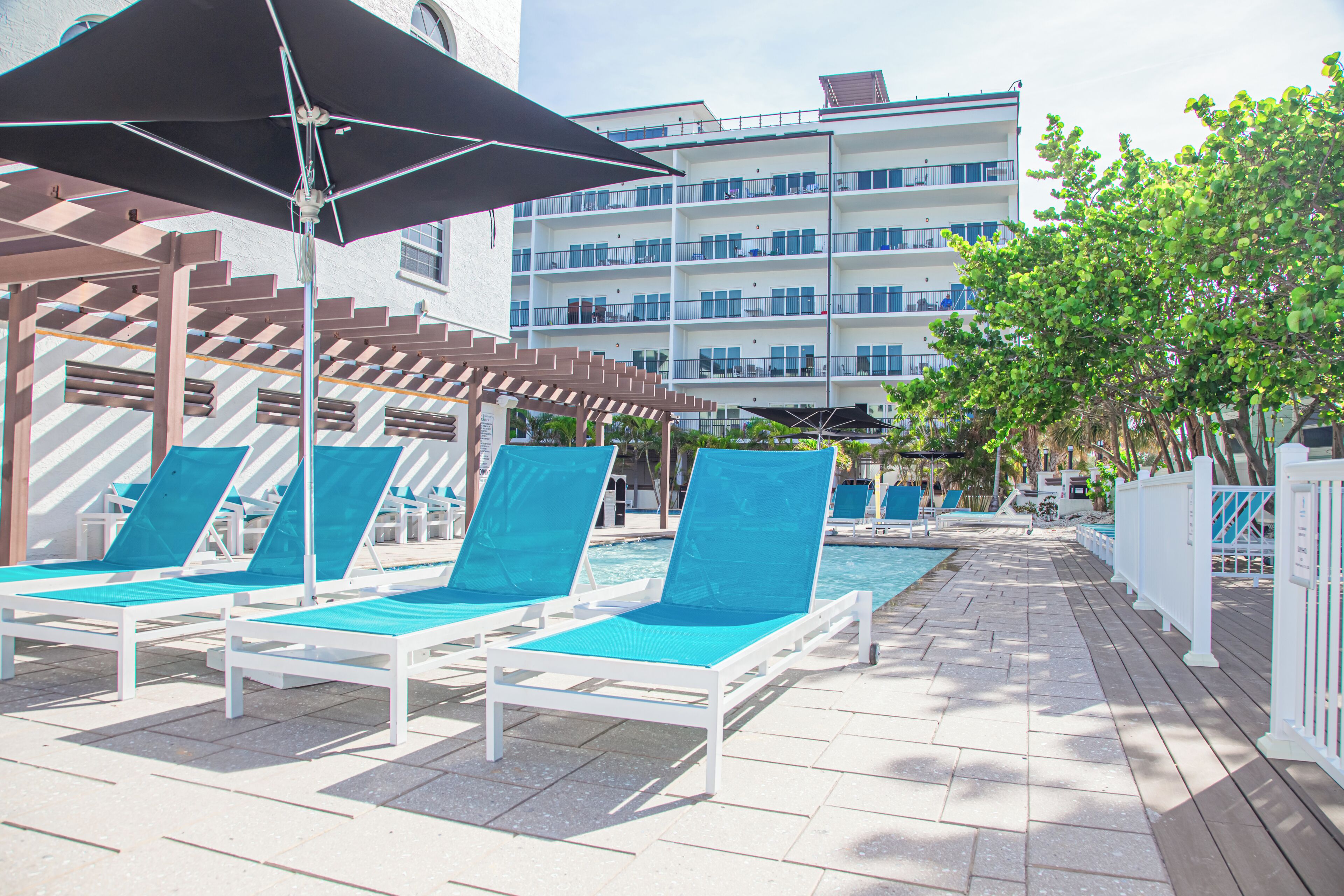 A poolside area with lounge chairs and umbrellas, a wooden pergola, and a multi-story building in the background.