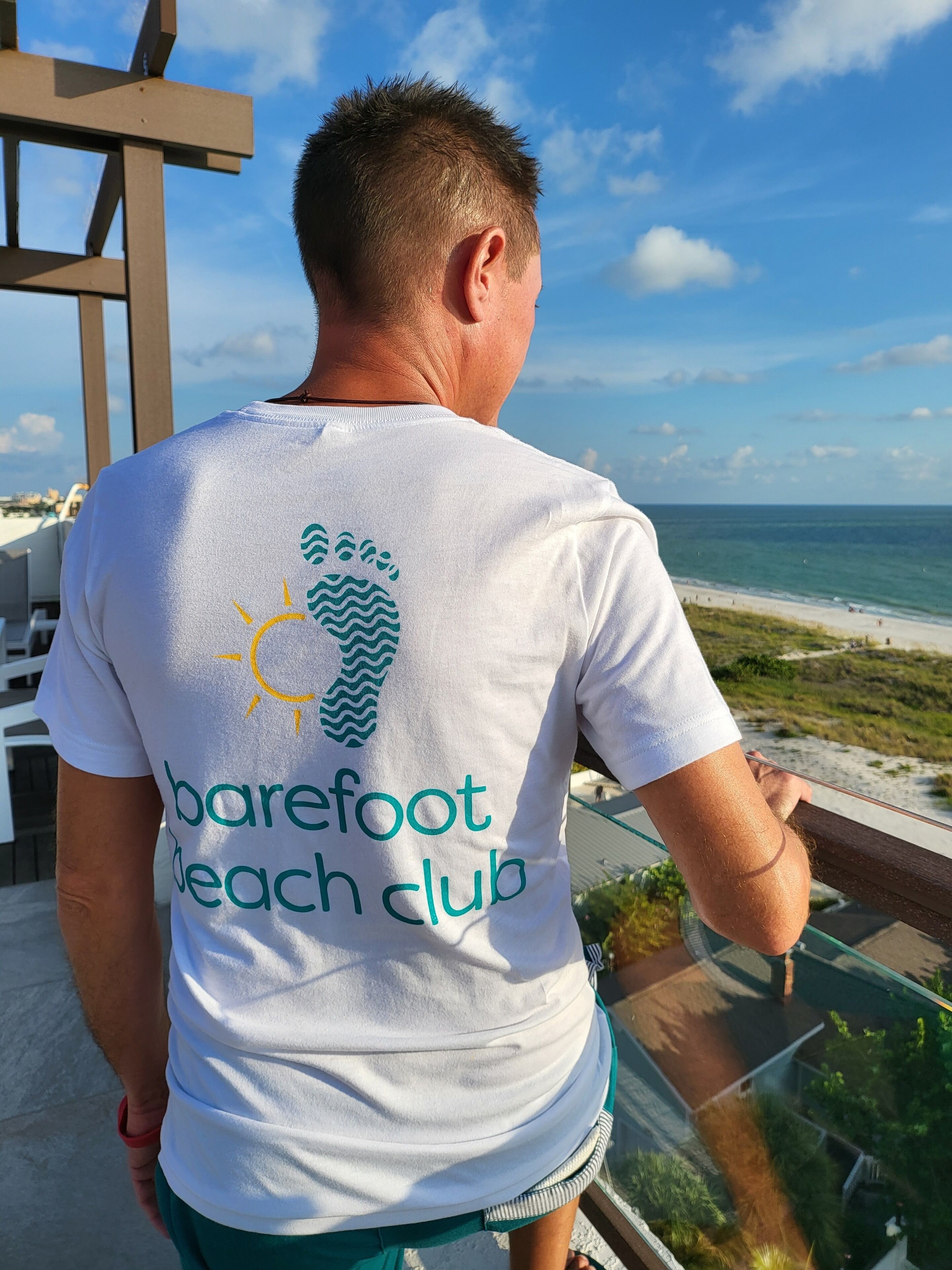 Eine Person in einem weißen T-Shirt mit dem Logo und Schriftzug eines Beach Clubs steht an einem Glasgeländer mit Blick auf einen Strand.