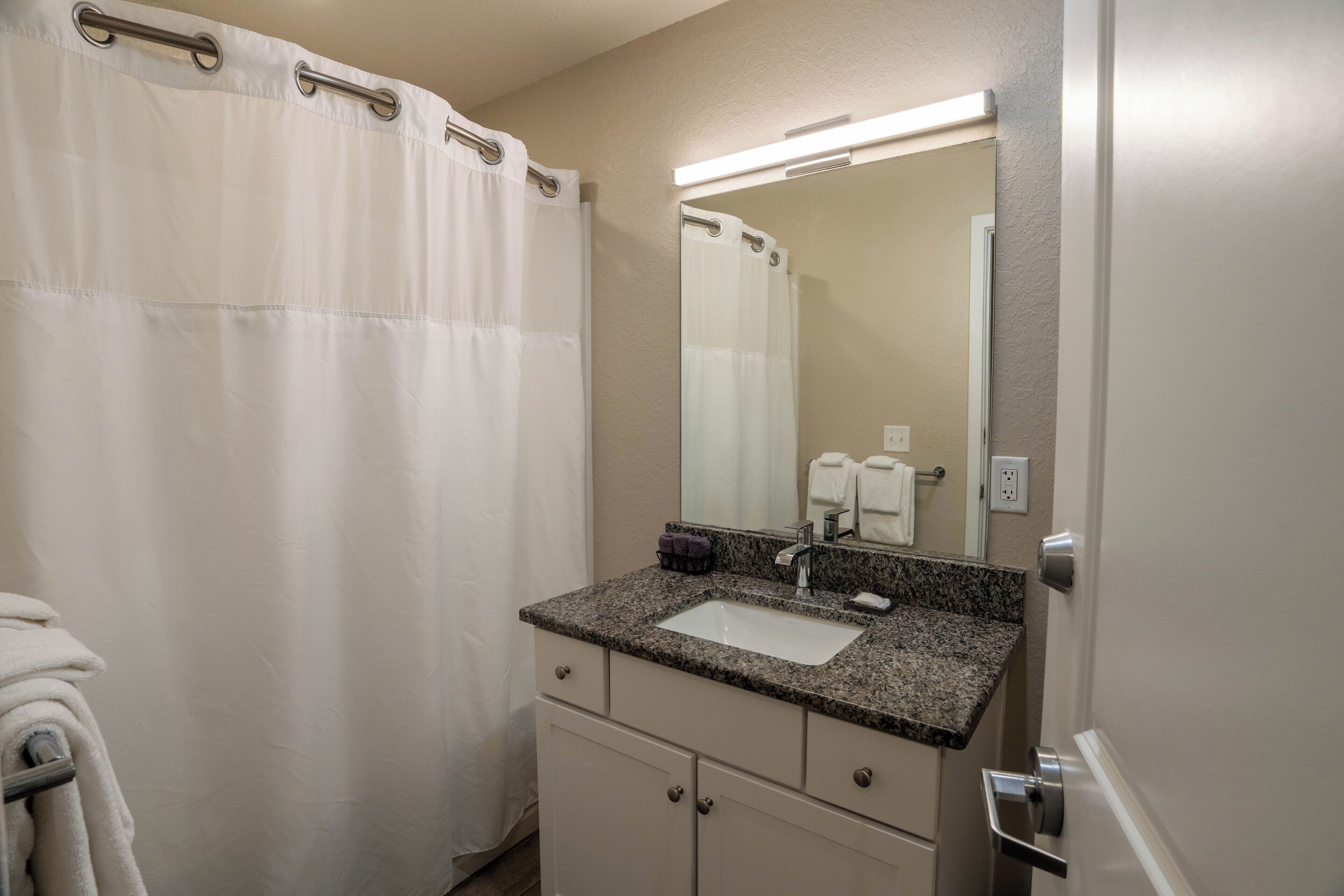 Une salle de bain avec un rideau de douche blanc, un lavabo doté d’un plan de travail en granit, un miroir et un porte-serviettes.