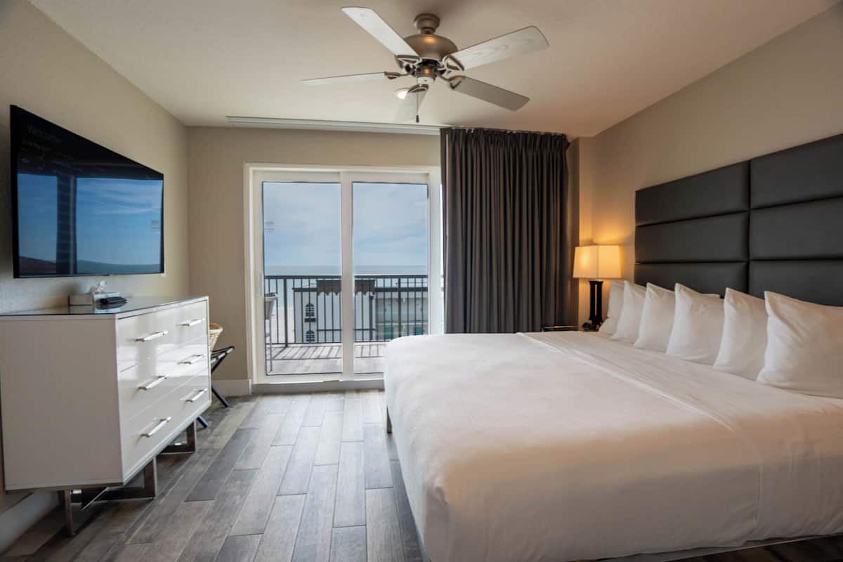 Une chambre d’hôtel avec un grand lit, un ventilateur de plafond, une table de chevet avec une lampe, une commode et une vue sur l’extérieur grâce à une porte coulissante.