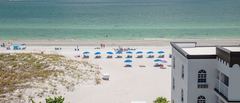 A beach with white sand, blue umbrellas, and a building with balconies.