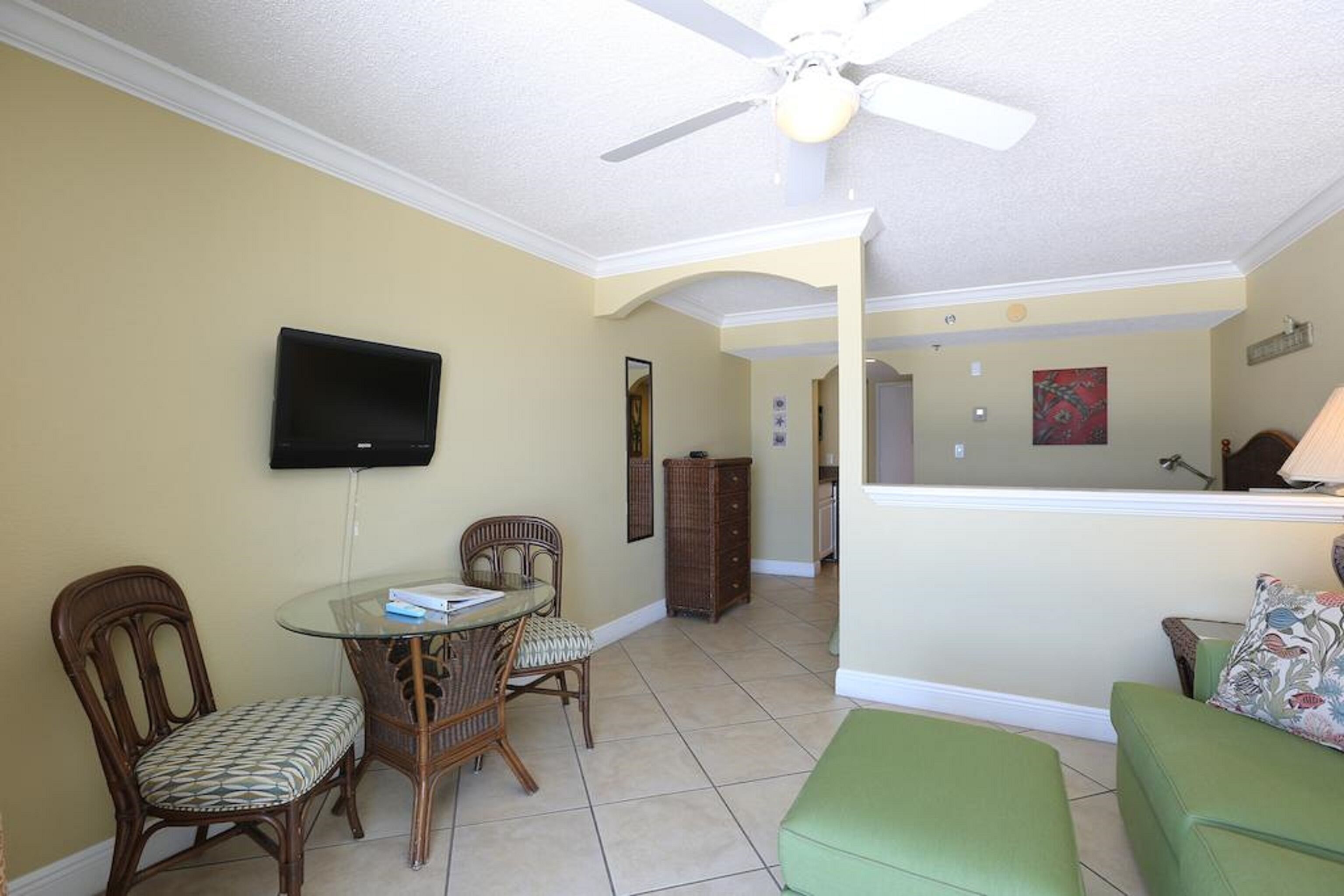A dining area with a glass table and wicker chairs, a ceiling fan, a television mounted on the wall, and a green sofa.