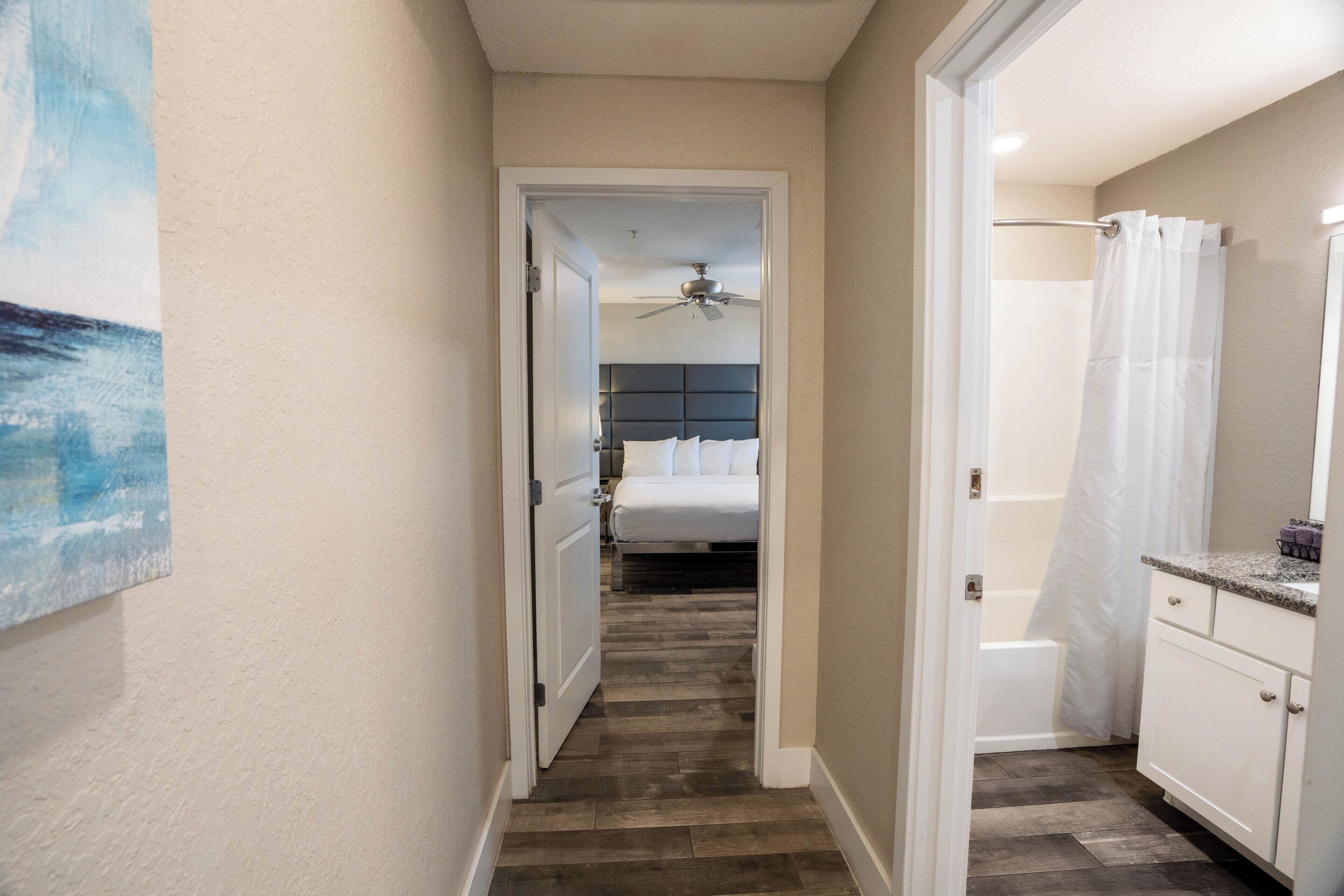 Un couloir étroit mène à une chambre à coucher équipée d’un lit, d’un ventilateur de plafond et d’une salle de bain avec un rideau de douche.
