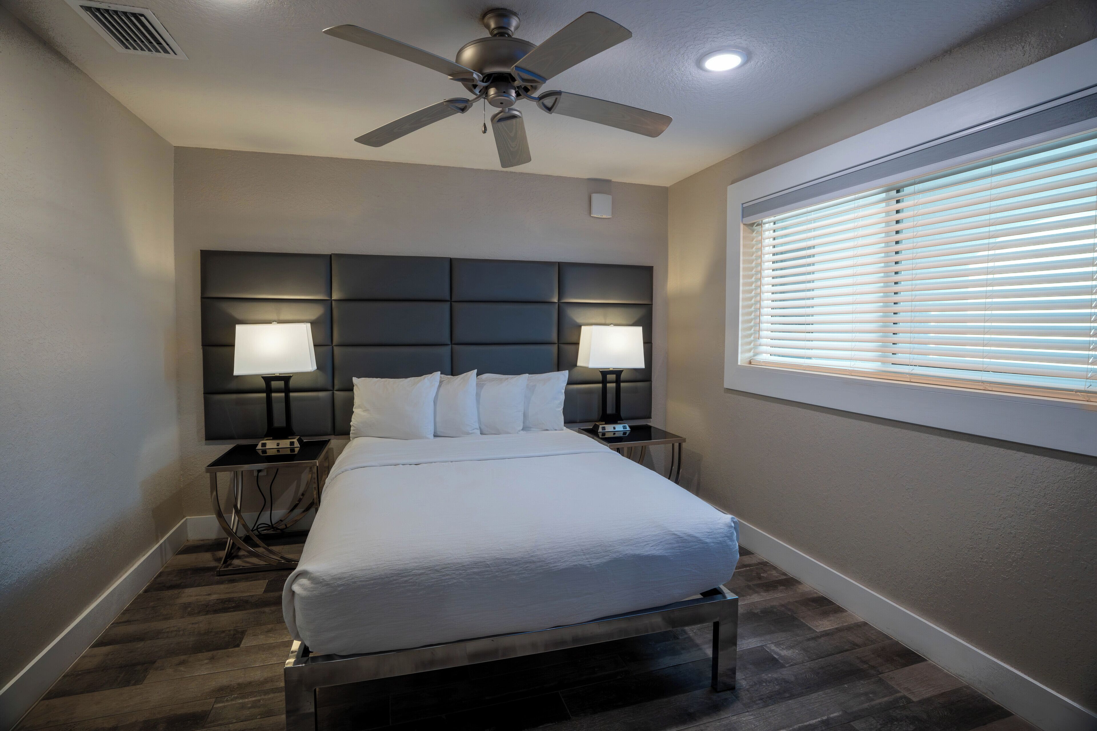 A hotel room with a large bed, two bedside tables with lamps, a ceiling fan, and a window with blinds.