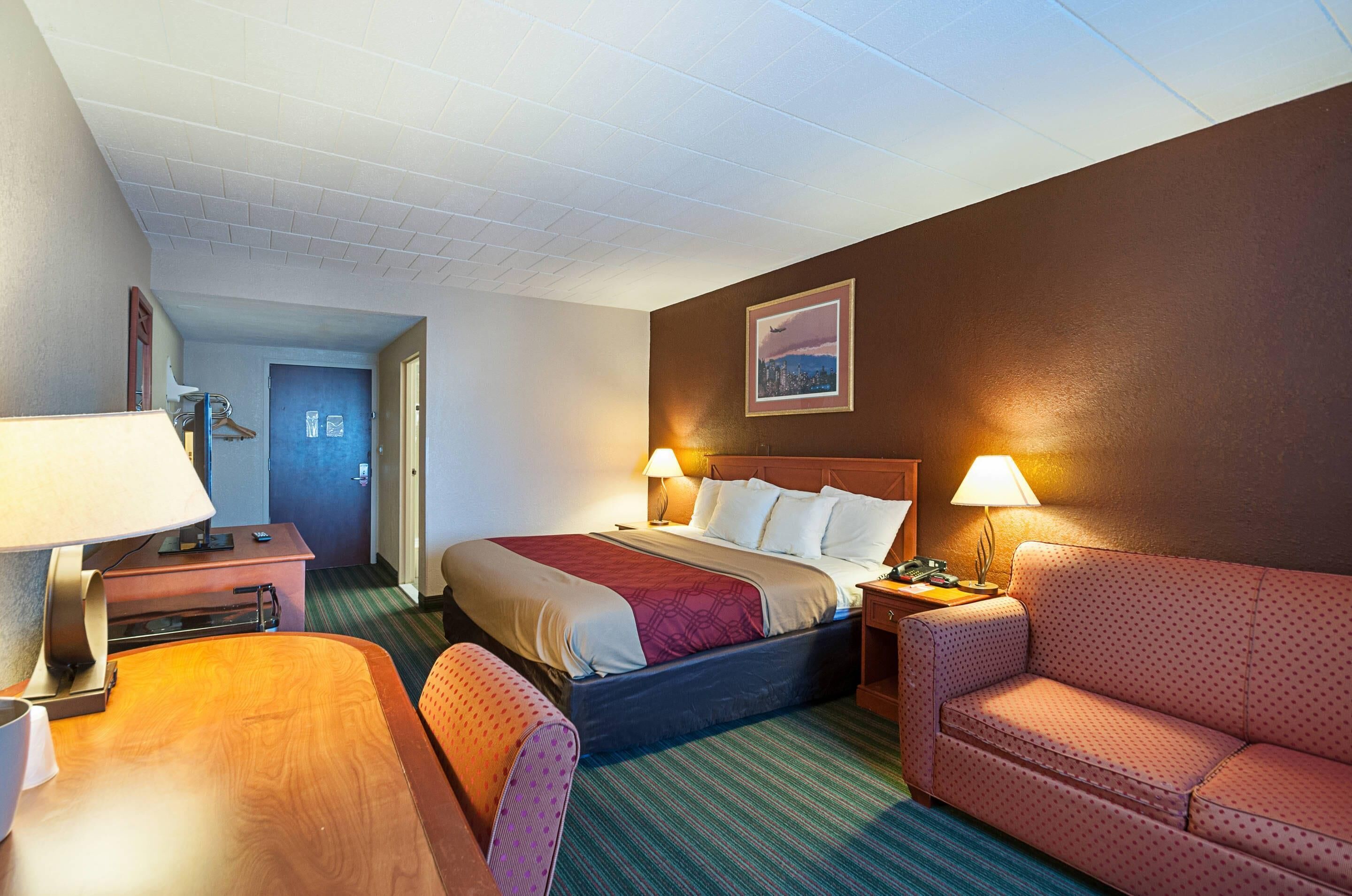 A hotel room with a bed, a desk, a chair, a sofa, and a lamp.
