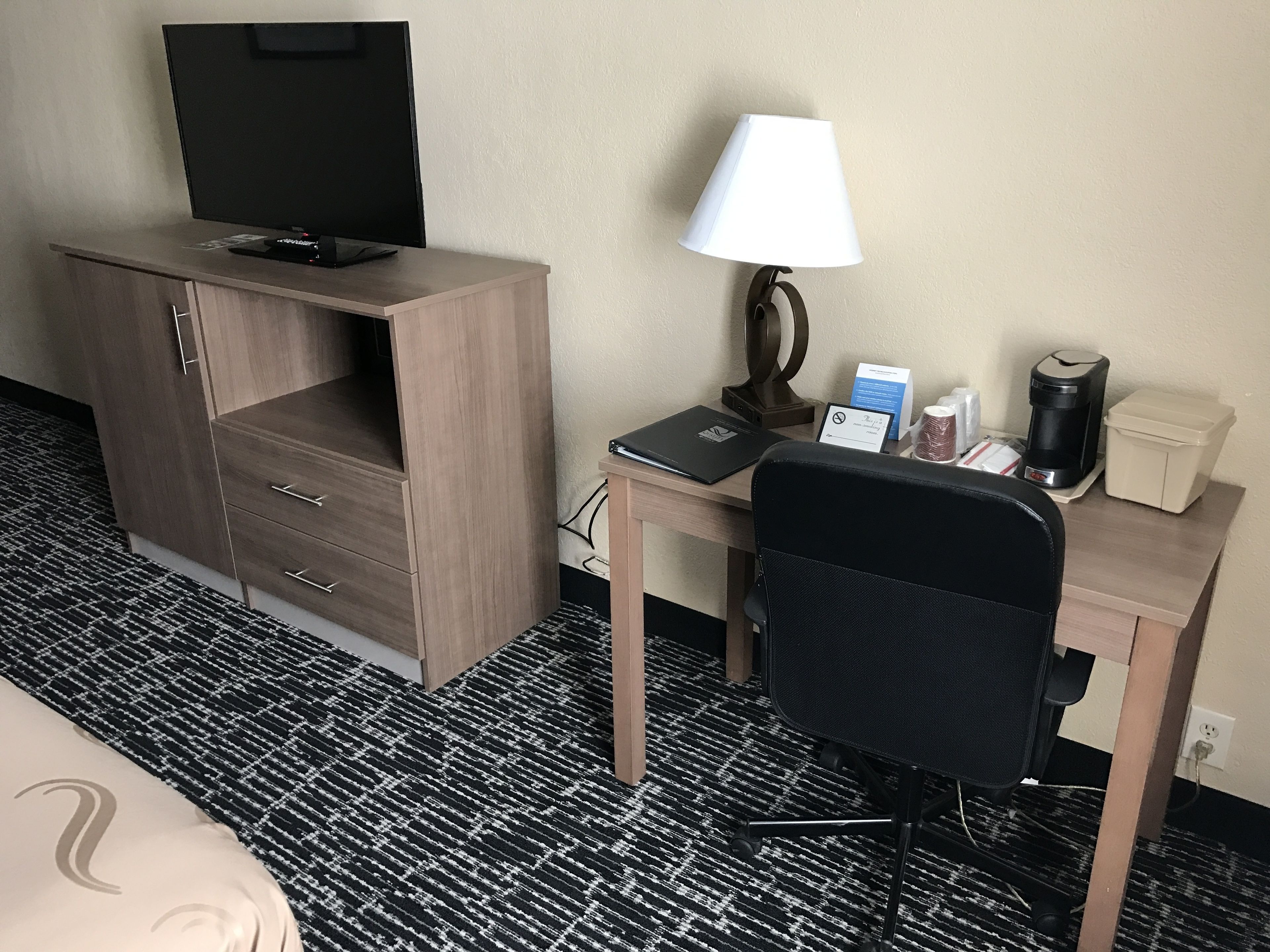 A hotel room with a wooden desk, a black office chair, a television, a lamp, a coffee maker, and a small table with a sign.